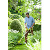 Man trimming garden plants with a yellow cordless grass trimmer, surrounded by greenery and bushes.